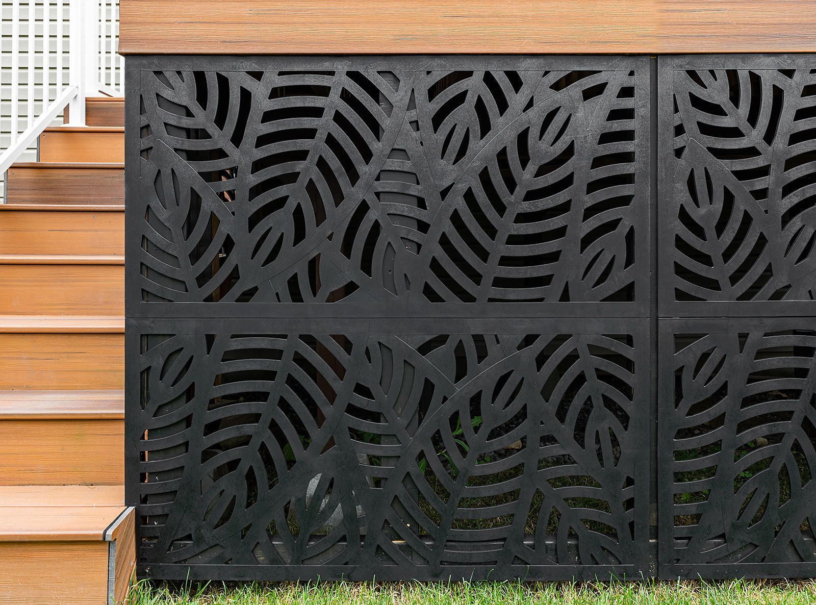 Panneaux décoratifs noirs à motifs de feuilles sous les marches d'une terrasse en bois, sur une zone gazonnée.