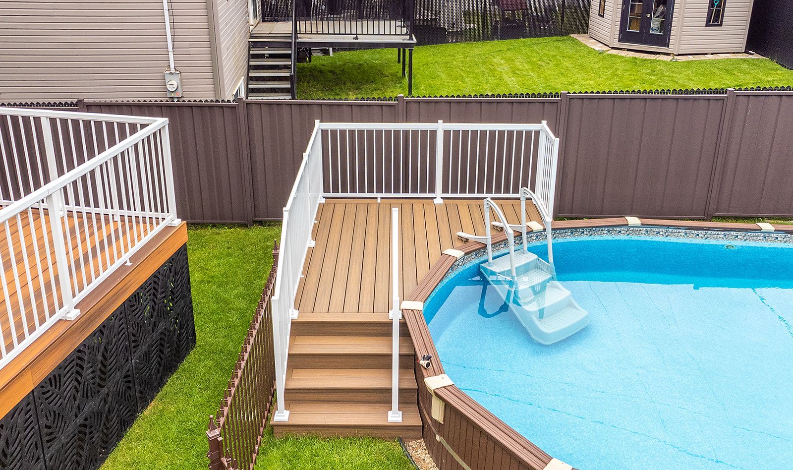 Terrasse avec rambarde blanche et marches menant à une piscine, entourée d'une clôture marron et de pelouse verte.