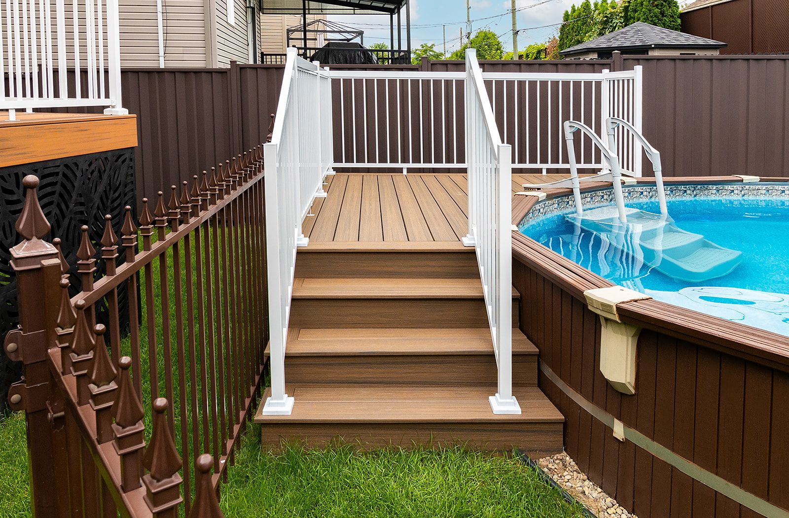 Des marches en bois avec des rambardes blanches mènent à une terrasse de piscine ; une clôture marron se trouve à gauche, une piscine bleue à droite.