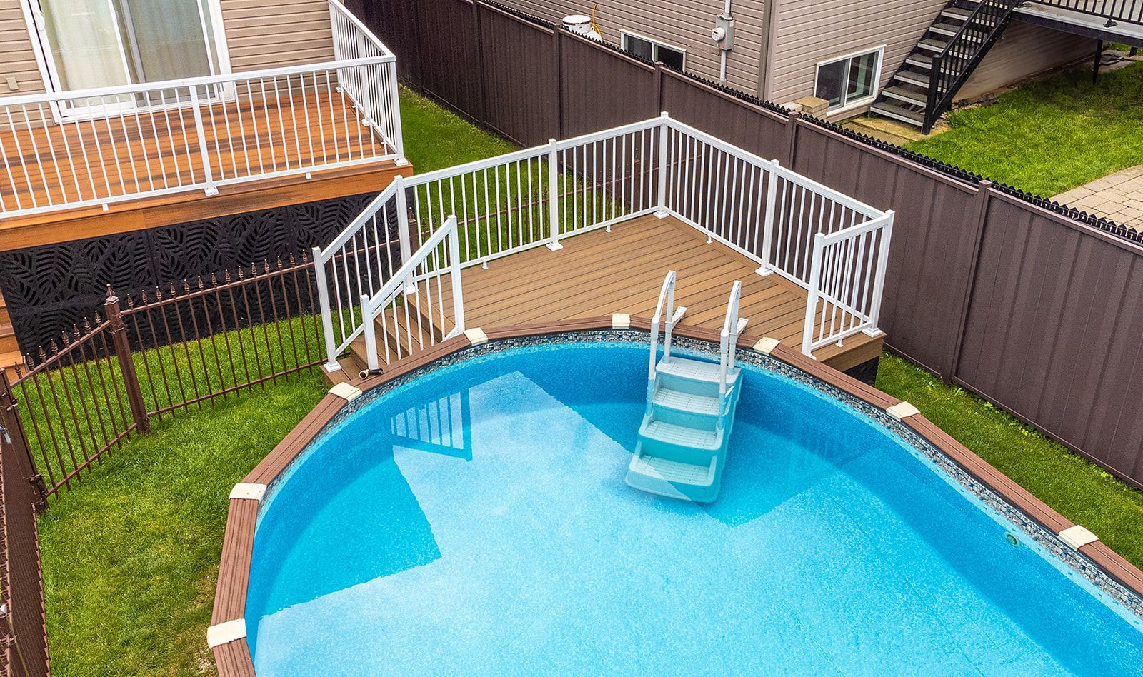 Piscine hors sol ovale avec escalier, entourée d'une terrasse en bois et d'une rambarde blanche. Clôture marron en arrière-plan.
