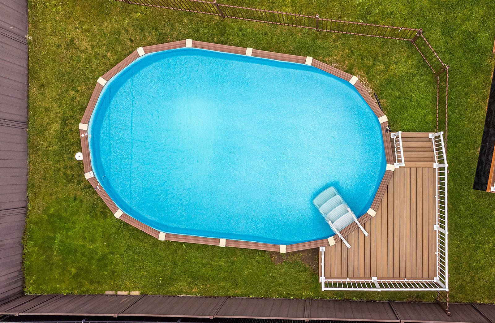 Vue aérienne d'une piscine ovale avec terrasse en bois et échelle, entourée de pelouse verte.