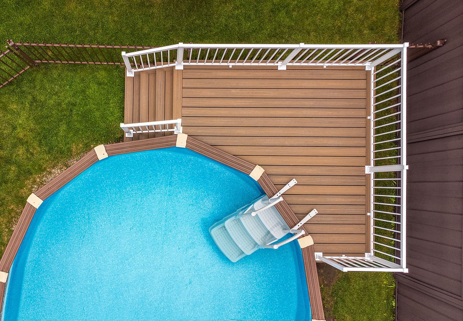 Vue aérienne d'une piscine hors sol avec terrasse en bois et rambarde blanche. Entourée de pelouse verte.