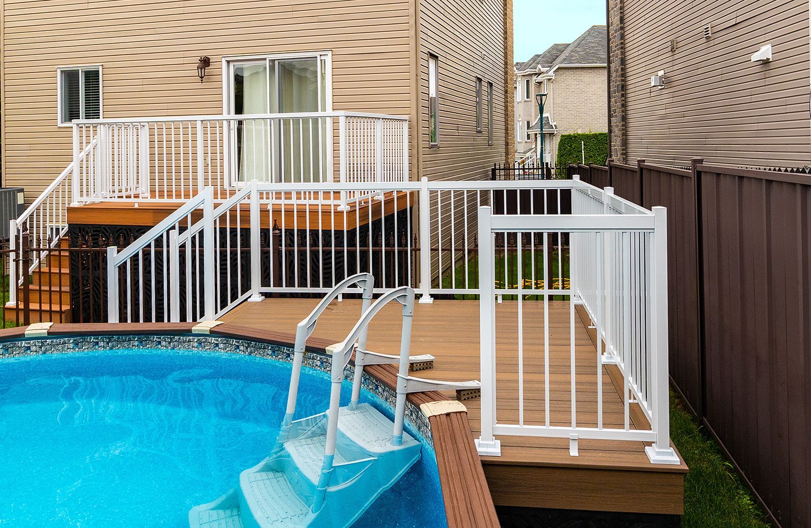 Une terrasse à balustrade blanche entoure une piscine hors sol. Un escalier en bois brun mène à une maison dotée d'une porte-fenêtre coulissante.