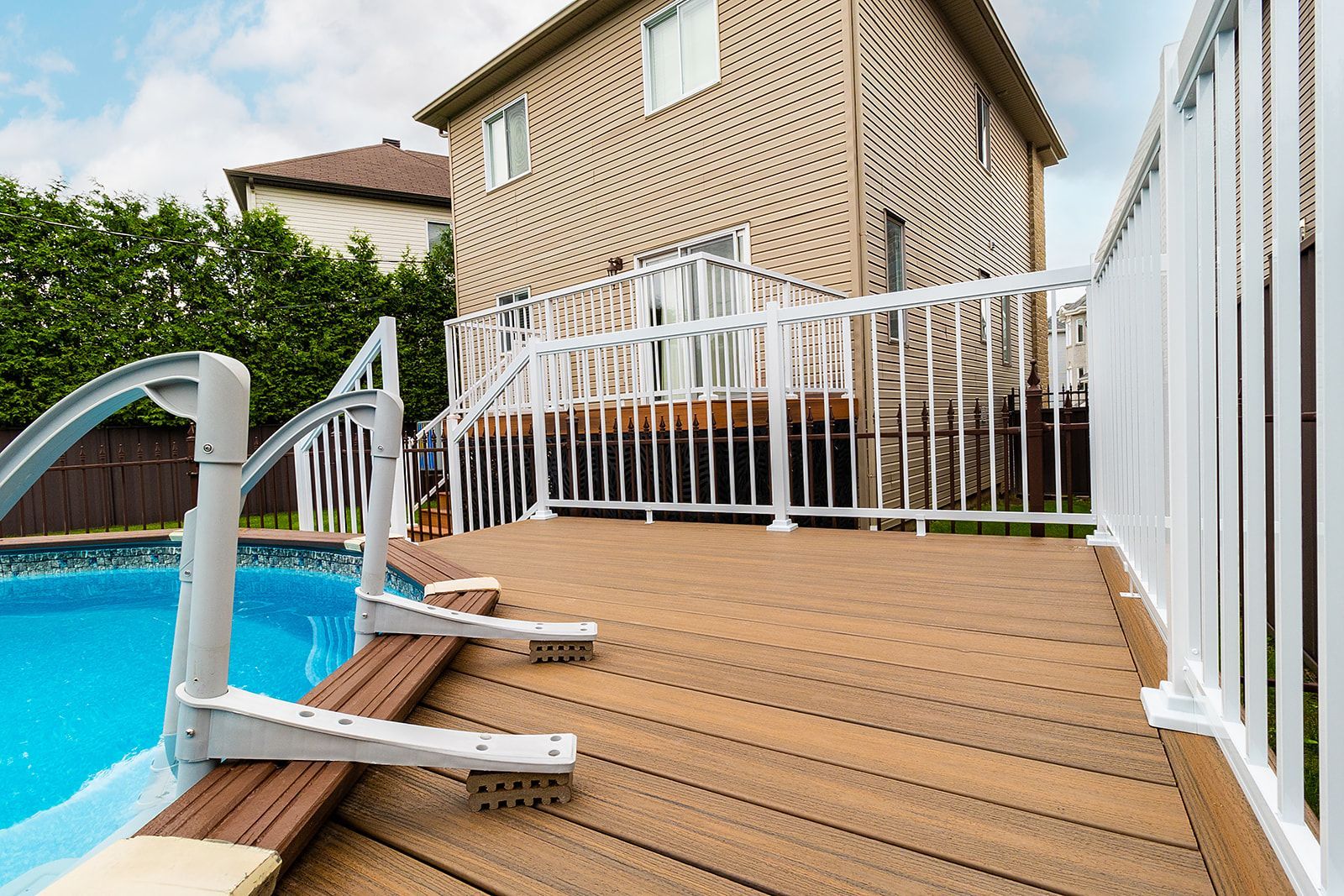Terrasse en composite avec rambarde blanche jouxtant une piscine et une maison en briques de deux étages. Échelle de piscine visible.