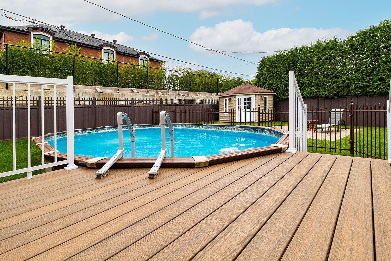 Piscine hors sol avec terrasse, entourée d'une clôture et d'une végétation luxuriante, journée ensoleillée.