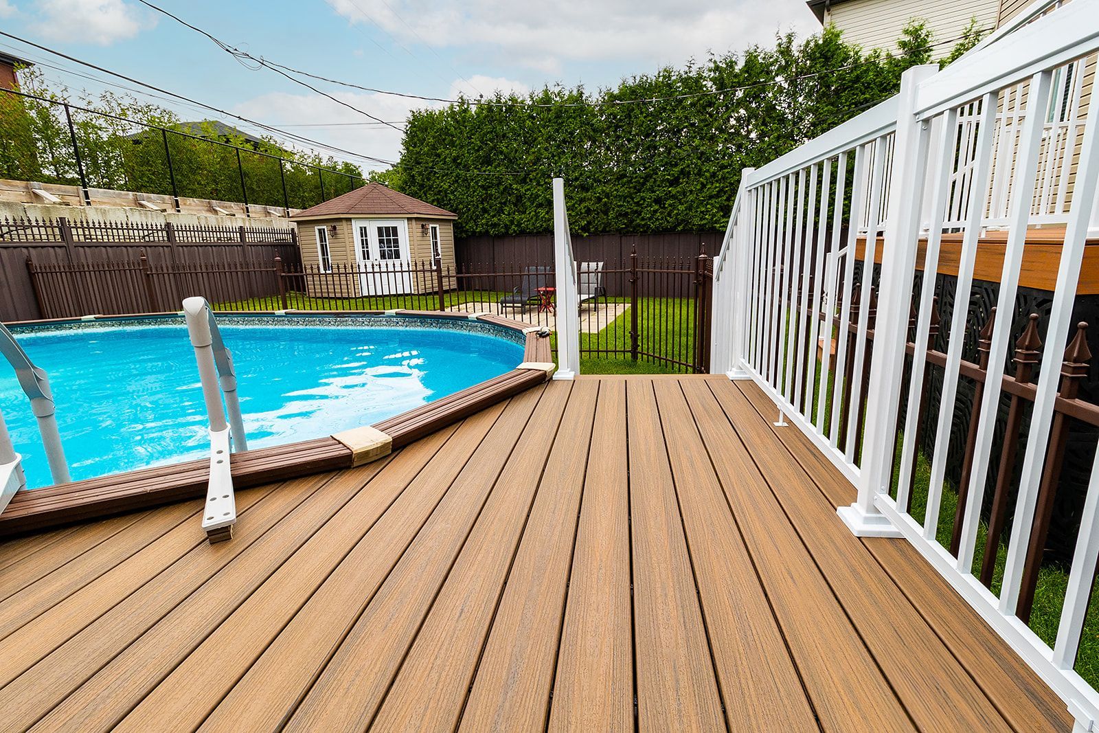 Terrasse en bois jouxtant une piscine avec une rambarde blanche, menant à un petit bâtiment en arrière-plan.