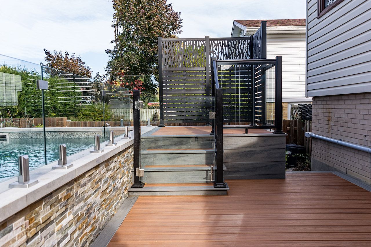 Terrasse en bois avec escalier menant à une zone surélevée entourée d'une clôture sombre et de balustrades en verre, près d'une piscine.