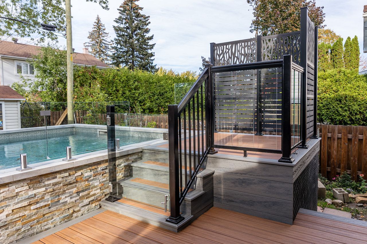 Cour arrière avec piscine et terrasse, balustrades noires, panneaux de verre, mur en pierre et terrasse en composite.