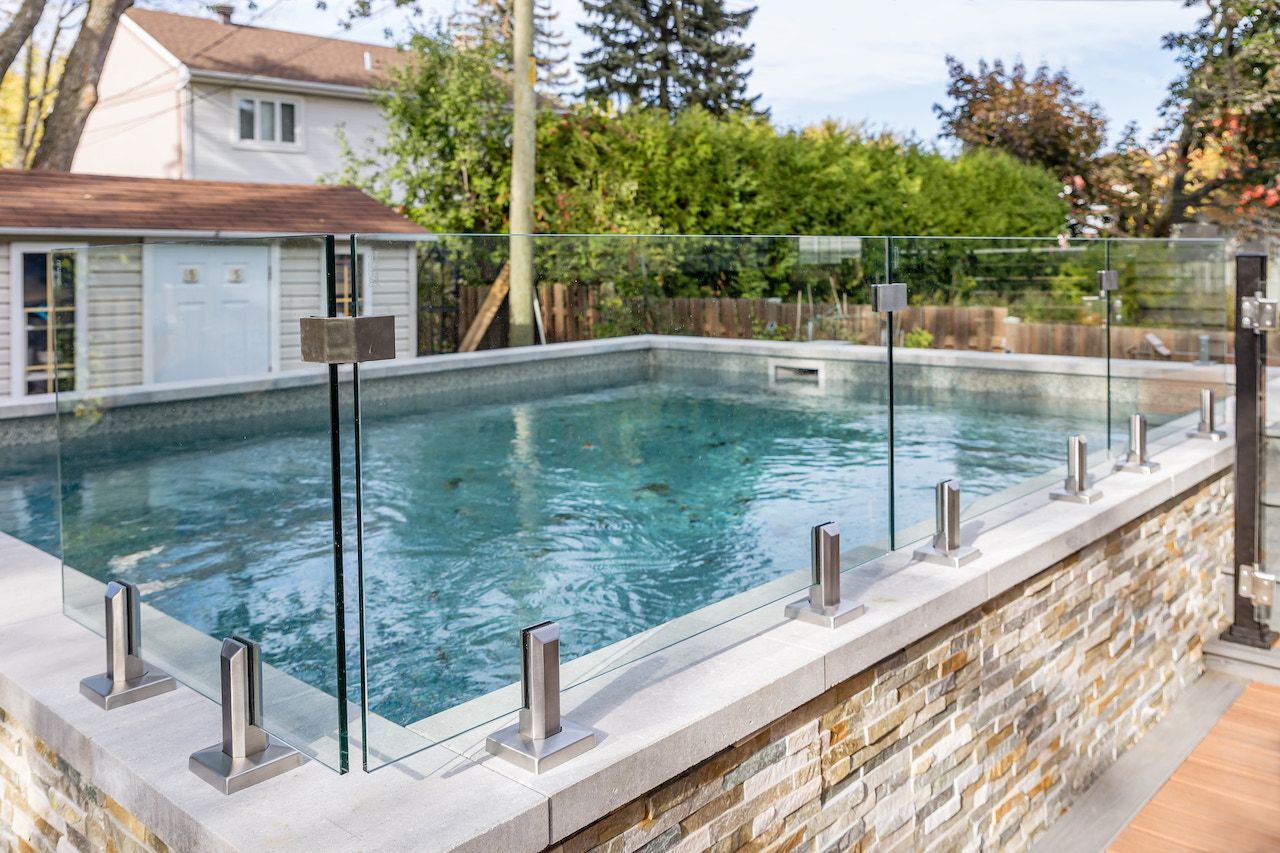 Piscine entourée d'une clôture de sécurité en verre, sur un rebord en pierre. Aménagement d'un jardin.