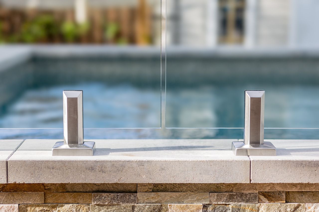 Clôture de piscine en verre avec poteaux en acier inoxydable, fixée sur un mur en pierre, piscine en arrière-plan.