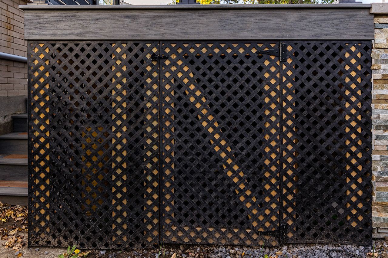Un treillis noir recouvre le dessous d'une terrasse, adossé à un mur de pierre.