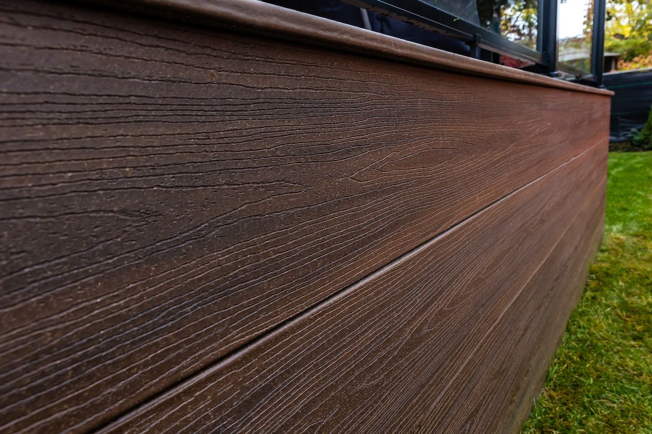 Gros plan sur une terrasse en composite marron adossée à un muret, à côté d'une pelouse verte.