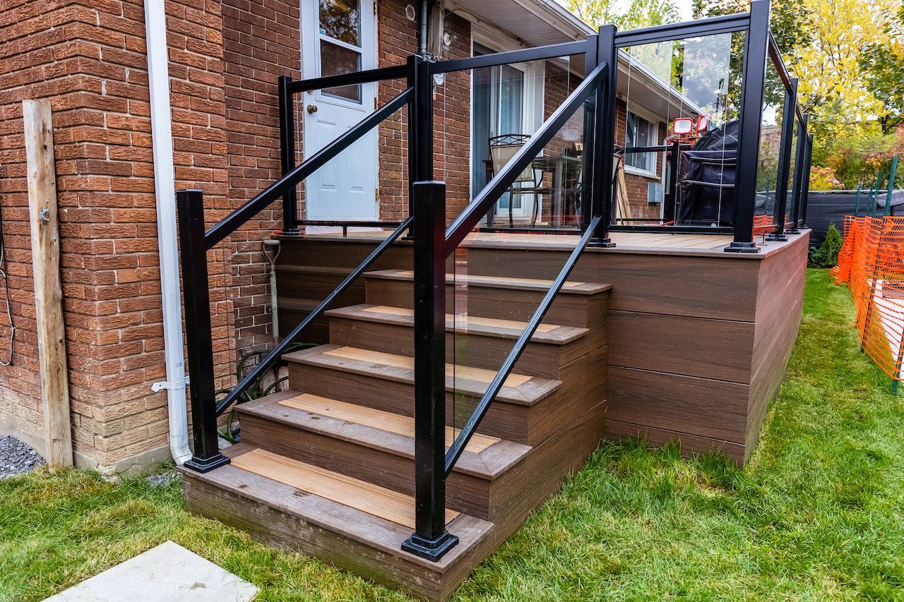 Une petite terrasse et un escalier avec une rambarde noire et des panneaux de verre, adossés à un bâtiment en briques.