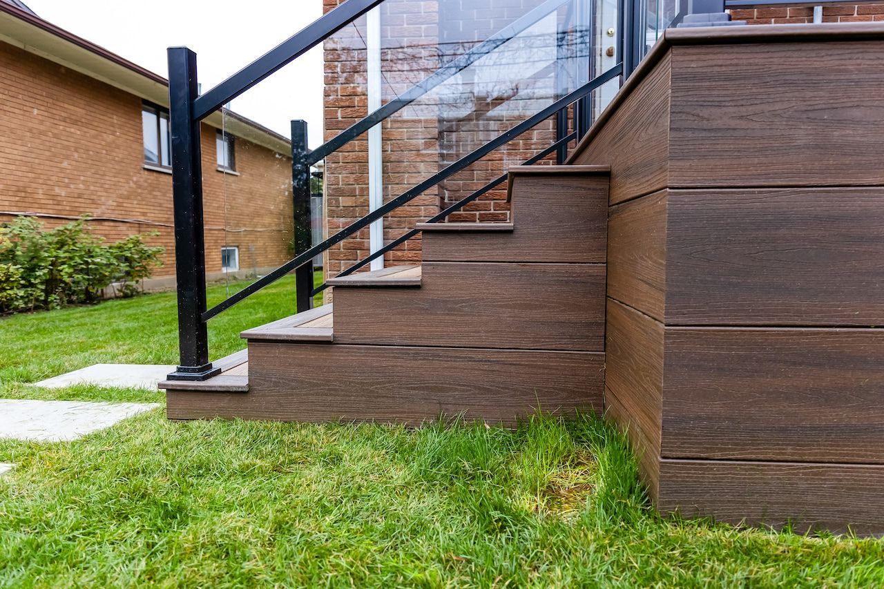 Terrasse en composite marron avec escalier et rambarde noire adossée à un bâtiment en briques, pelouse verte.