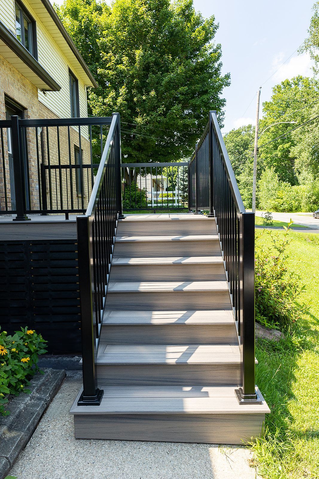 Un escalier en bois avec une rampe noire mène à une terrasse, près d'une maison et d'arbres.