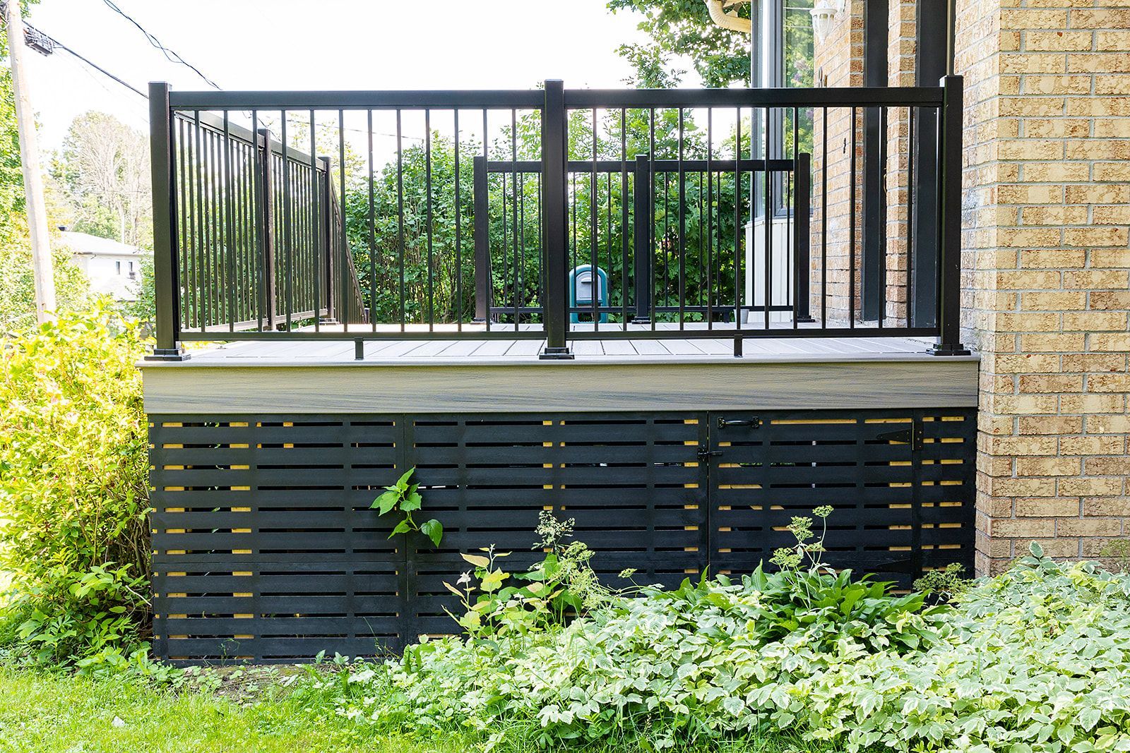 Terrasse noire avec rambarde assortie, adossée à un mur de briques et à de la végétation.