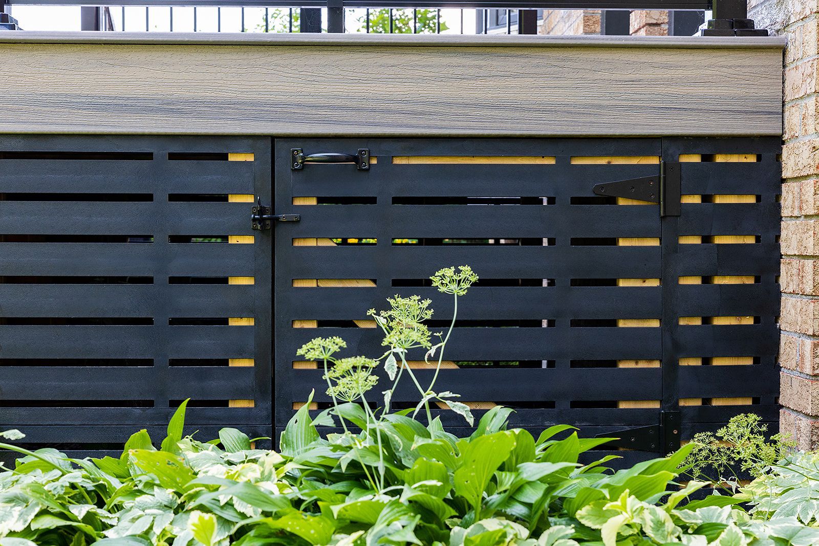 Portail à lattes noires, faisant partie d'une terrasse, avec des plantes devant.