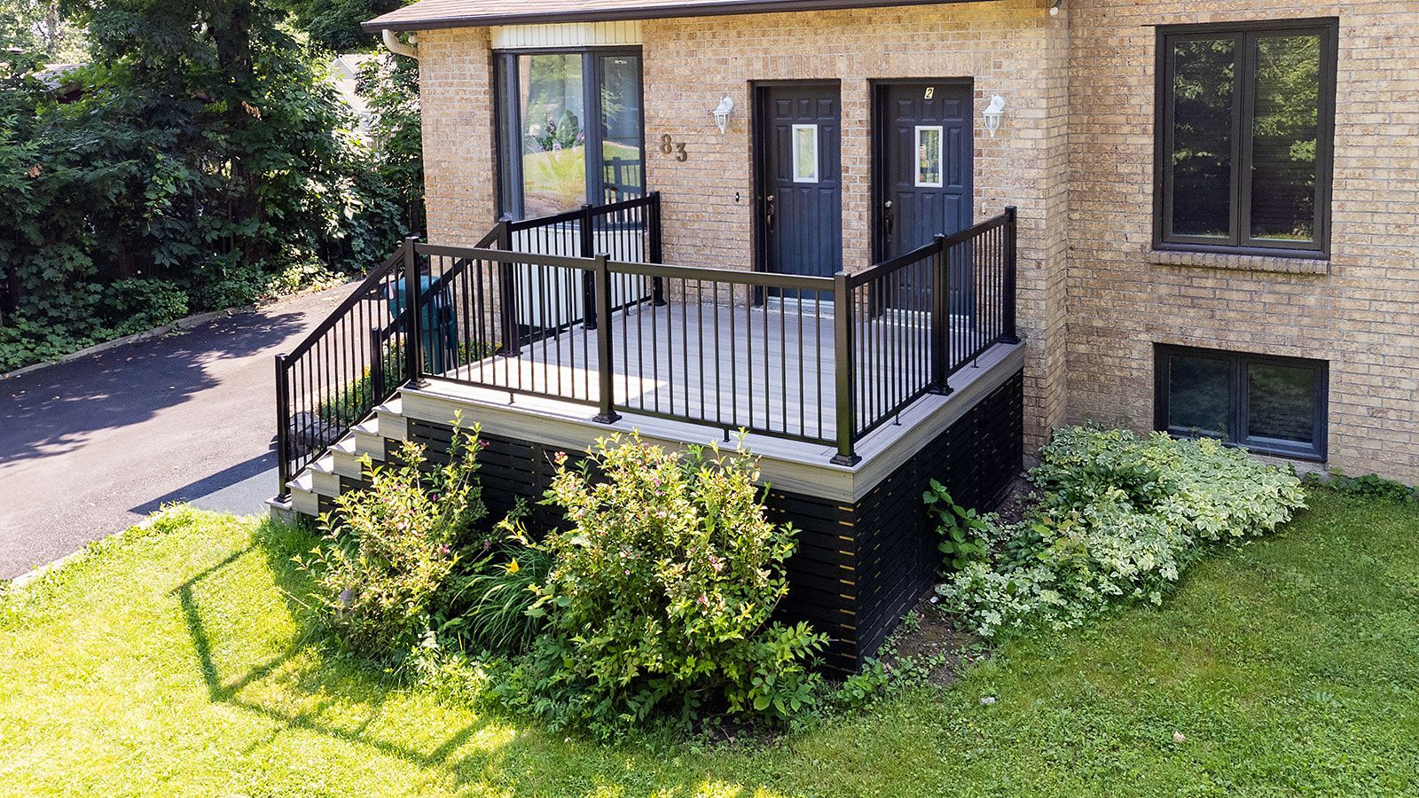 Extérieur d'un bâtiment en briques avec une terrasse et des balustrades noires, deux portes et de la verdure.
