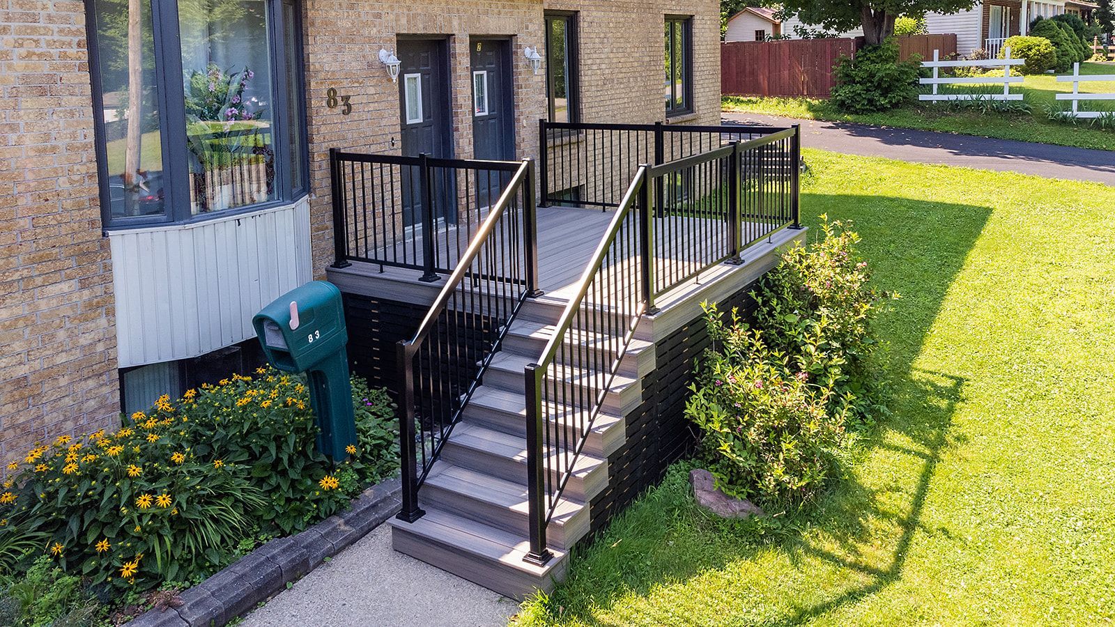 Entrée principale avec rambarde noire, escalier et terrasse. Pelouse verte, boîte aux lettres et bâtiment en briques.