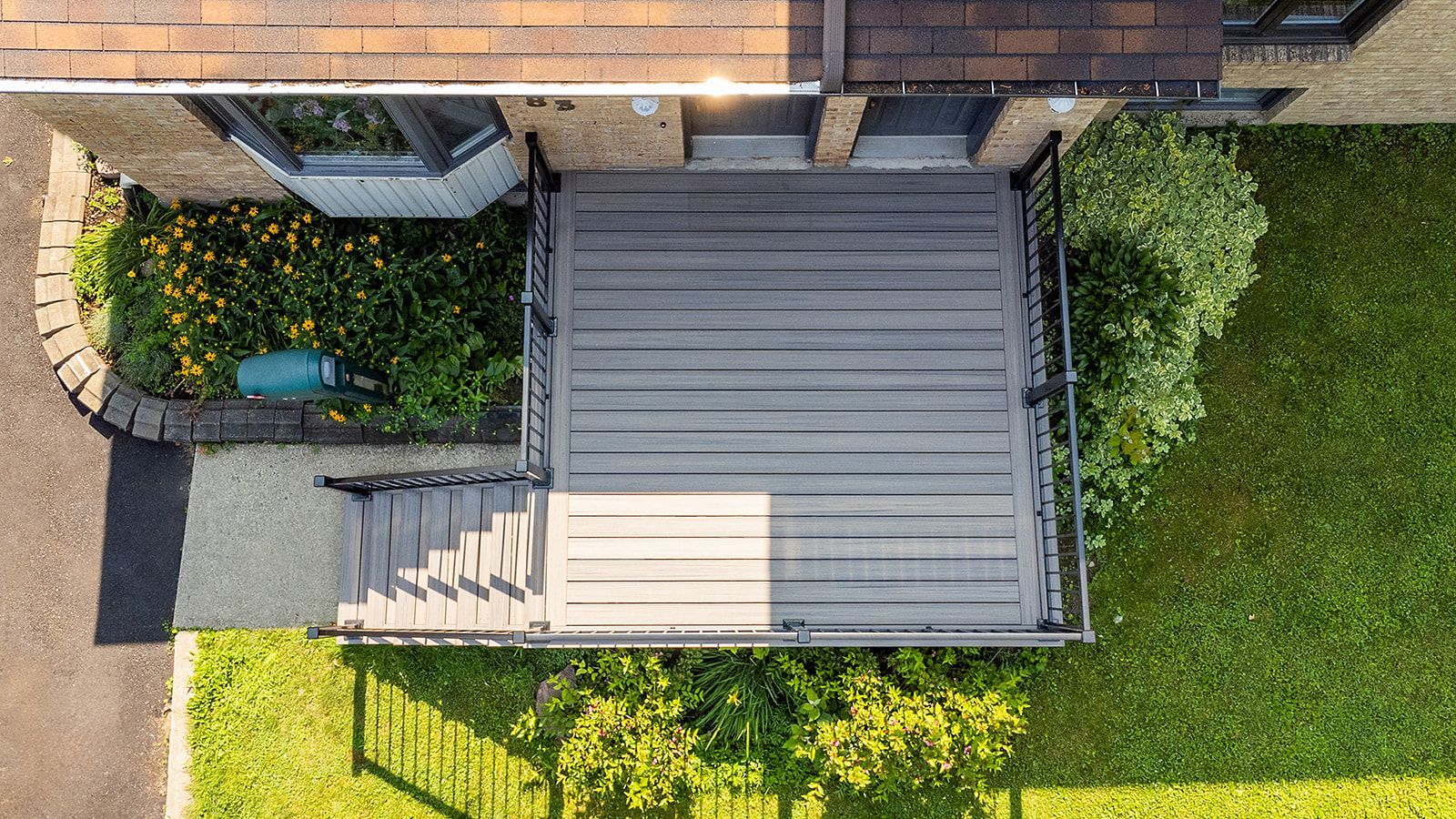 Vue aérienne d'une terrasse grise avec un escalier menant à une maison. Une pelouse verte agrémentée de quelques buissons et fleurs l'entoure.