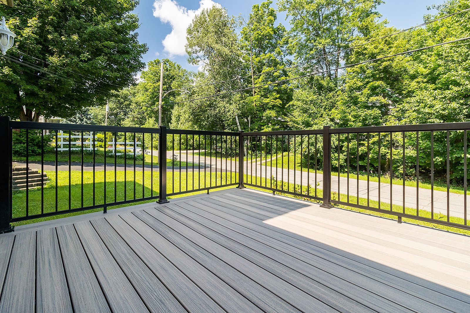 Terrasse en bois avec rambarde noire, donnant sur un jardin gazonné et arboré sous un ciel bleu.