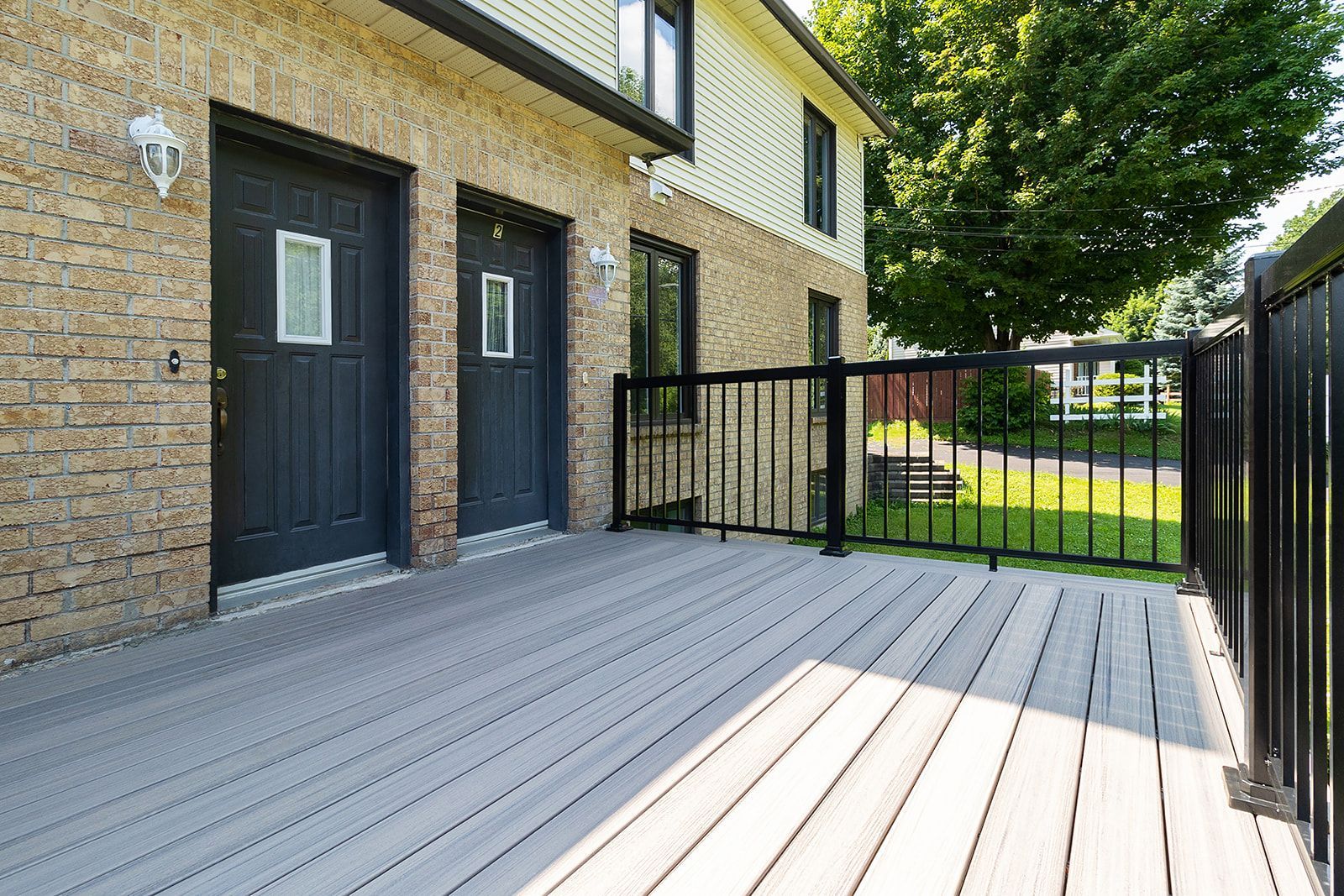 Terrasse avec planches grises, rambarde noire et deux portes noires sur une maison en briques.
