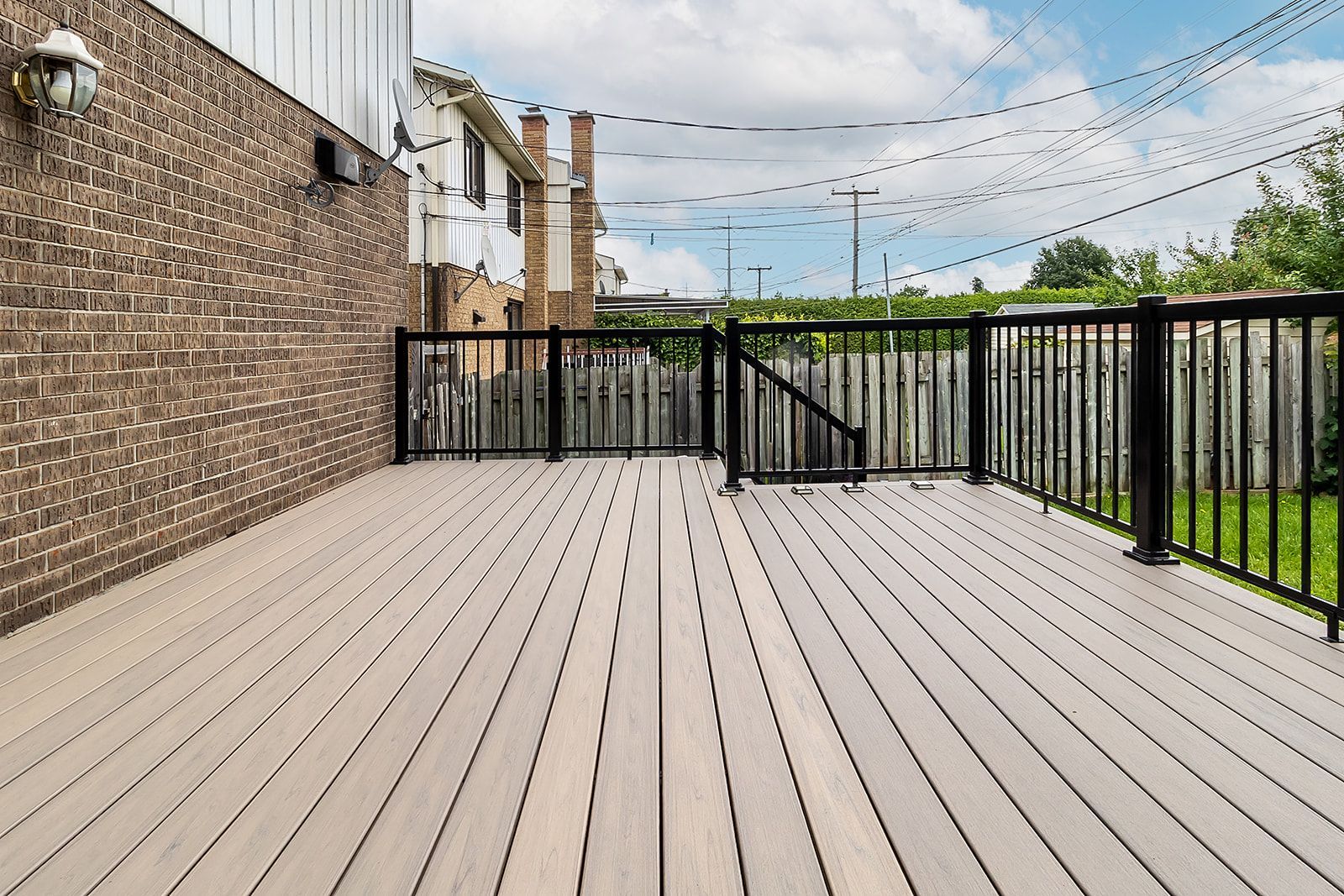 Terrasse en bois avec rambarde noire, jouxtant un mur de briques et une clôture, guirlandes lumineuses au-dessus et pelouse verte.