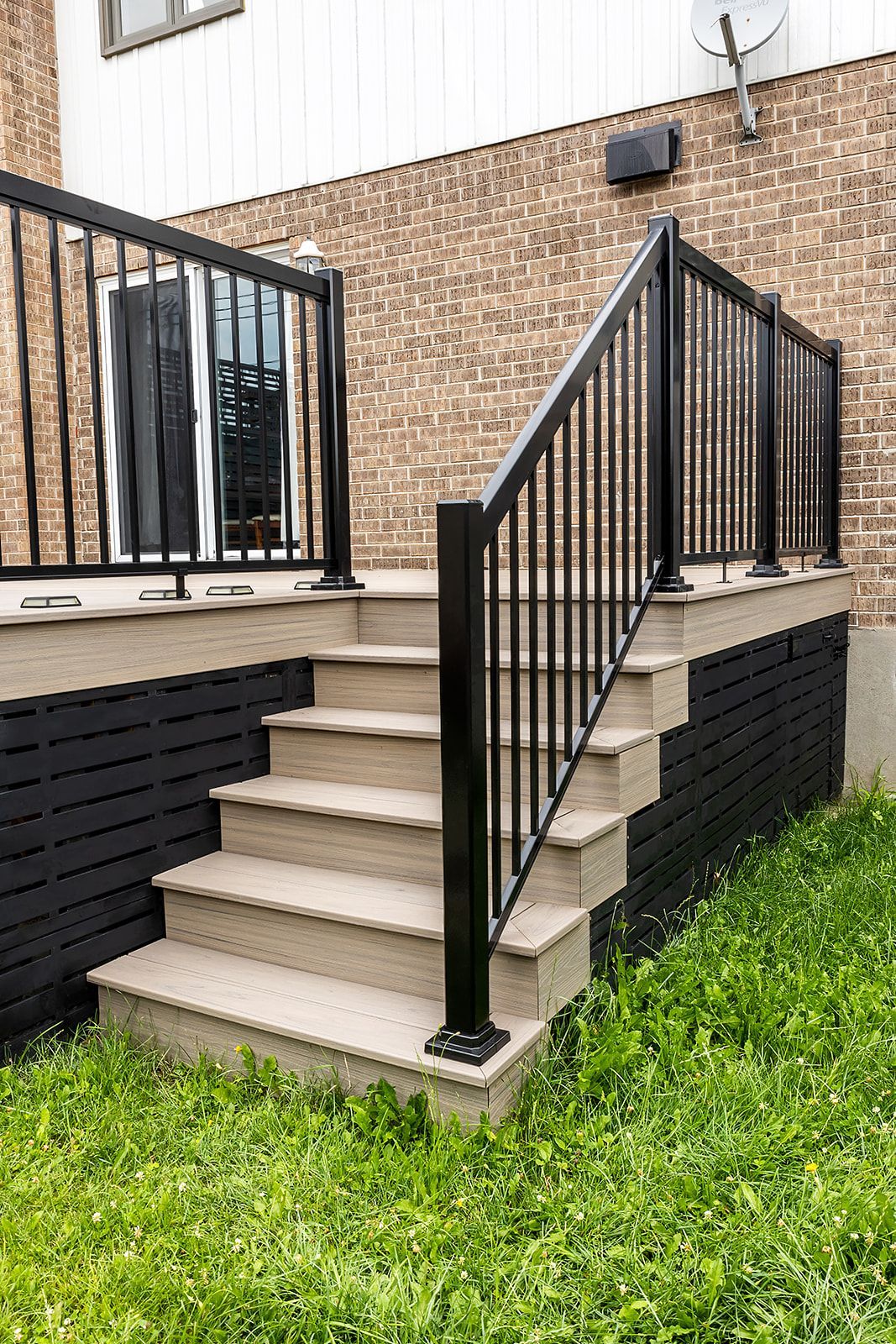 Escalier extérieur avec rampes noires et terrasse en composite, fond en briques et pelouse.