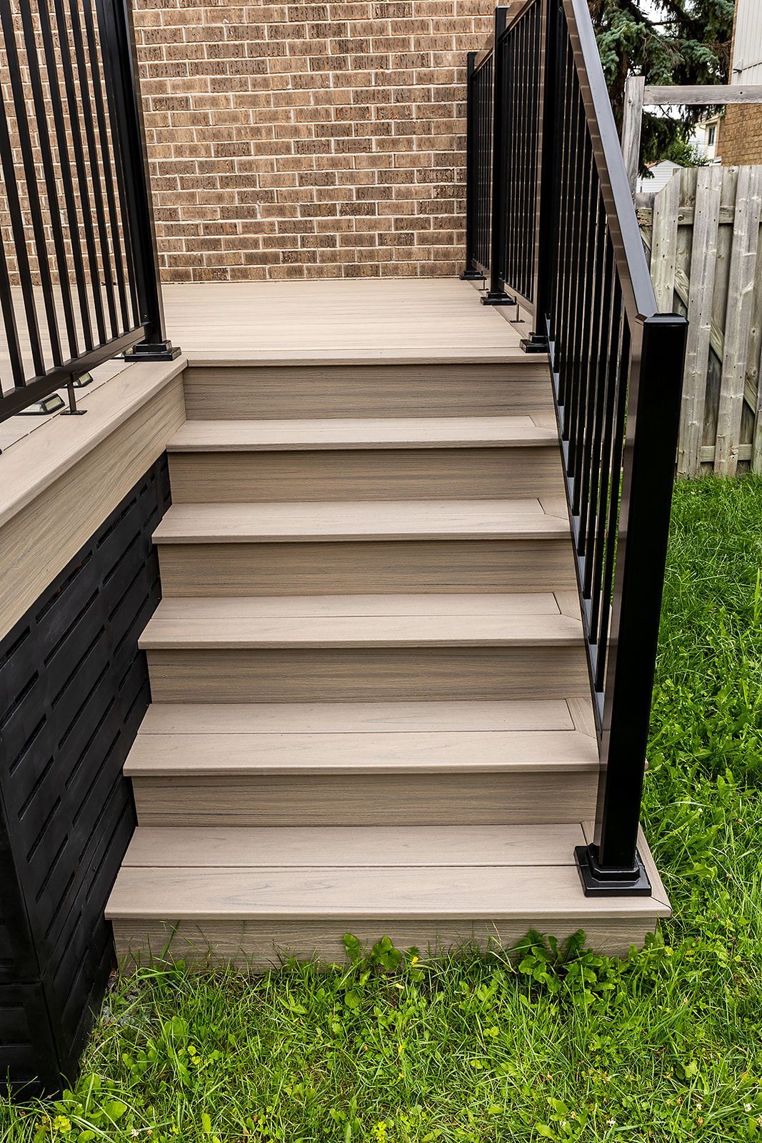 Escalier de terrasse en composite avec rambarde noire, adossé à un mur de briques et à une pelouse verte.