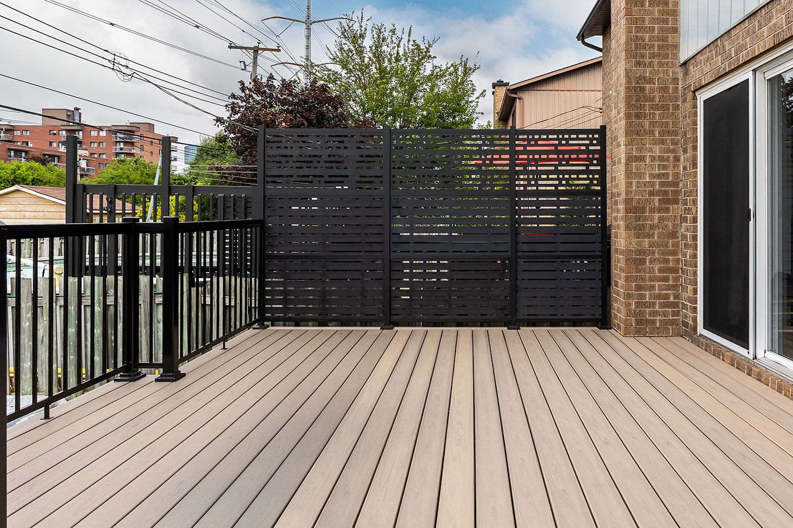 Une terrasse en bois avec une rambarde en métal noir et une clôture décorative pour préserver l'intimité. Façade de la maison et lignes électriques en arrière-plan.