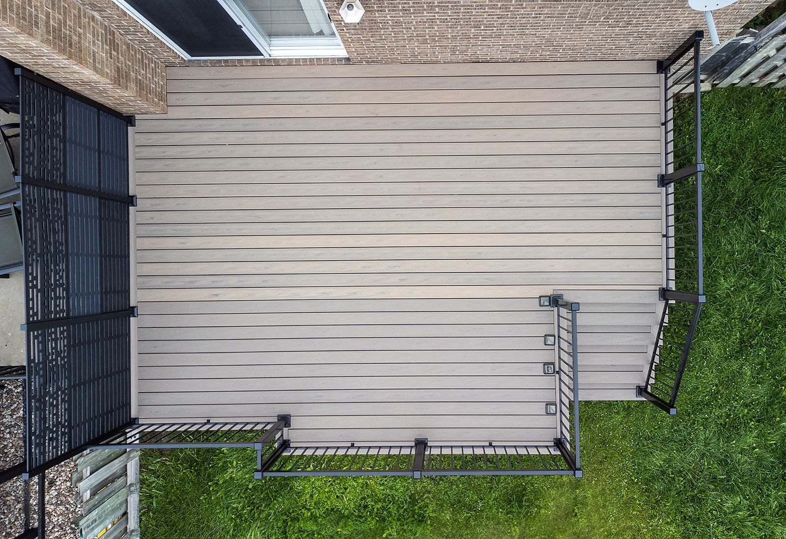 Vue aérienne d'une terrasse en composite avec des rambardes noires, jouxtant un mur de briques et une pelouse verte.