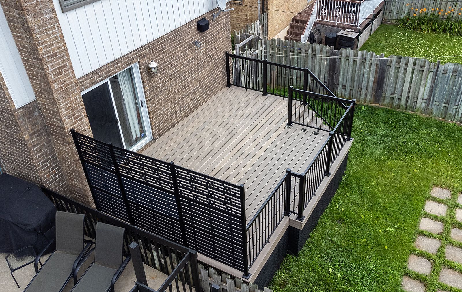 Une terrasse à plusieurs niveaux avec rambarde noire et brise-vue, située à l'extérieur d'une maison en briques et donnant sur un jardin gazonné.