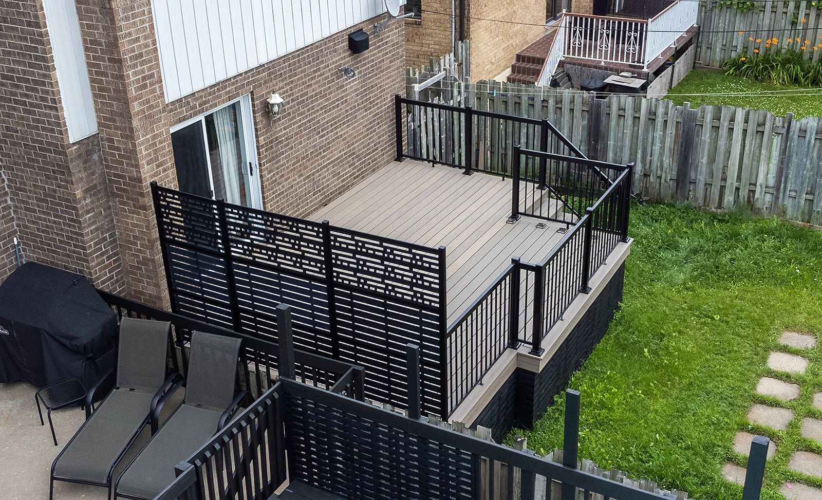 Terrasse arrière avec balustrade noire et brise-vue ; adjacente à une maison en briques et à une pelouse.