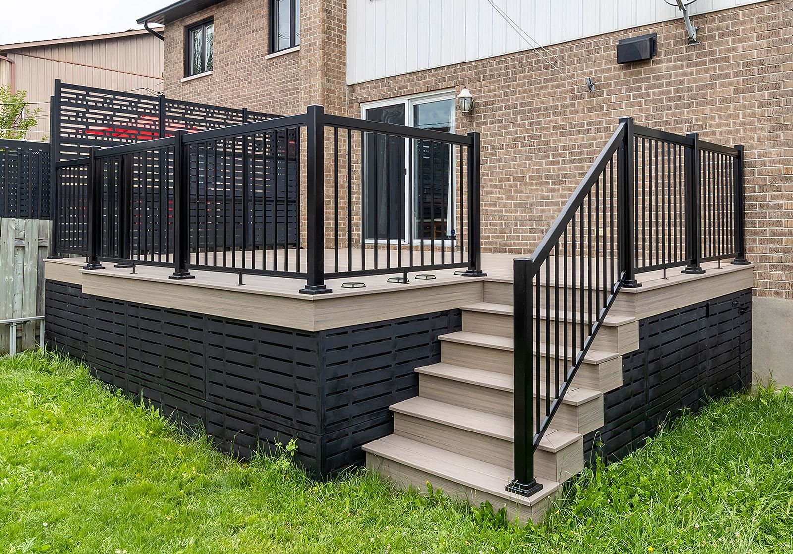 Terrasse noire avec escalier, rambarde et pergola adossée à un mur de briques. Entourée de pelouse verte.