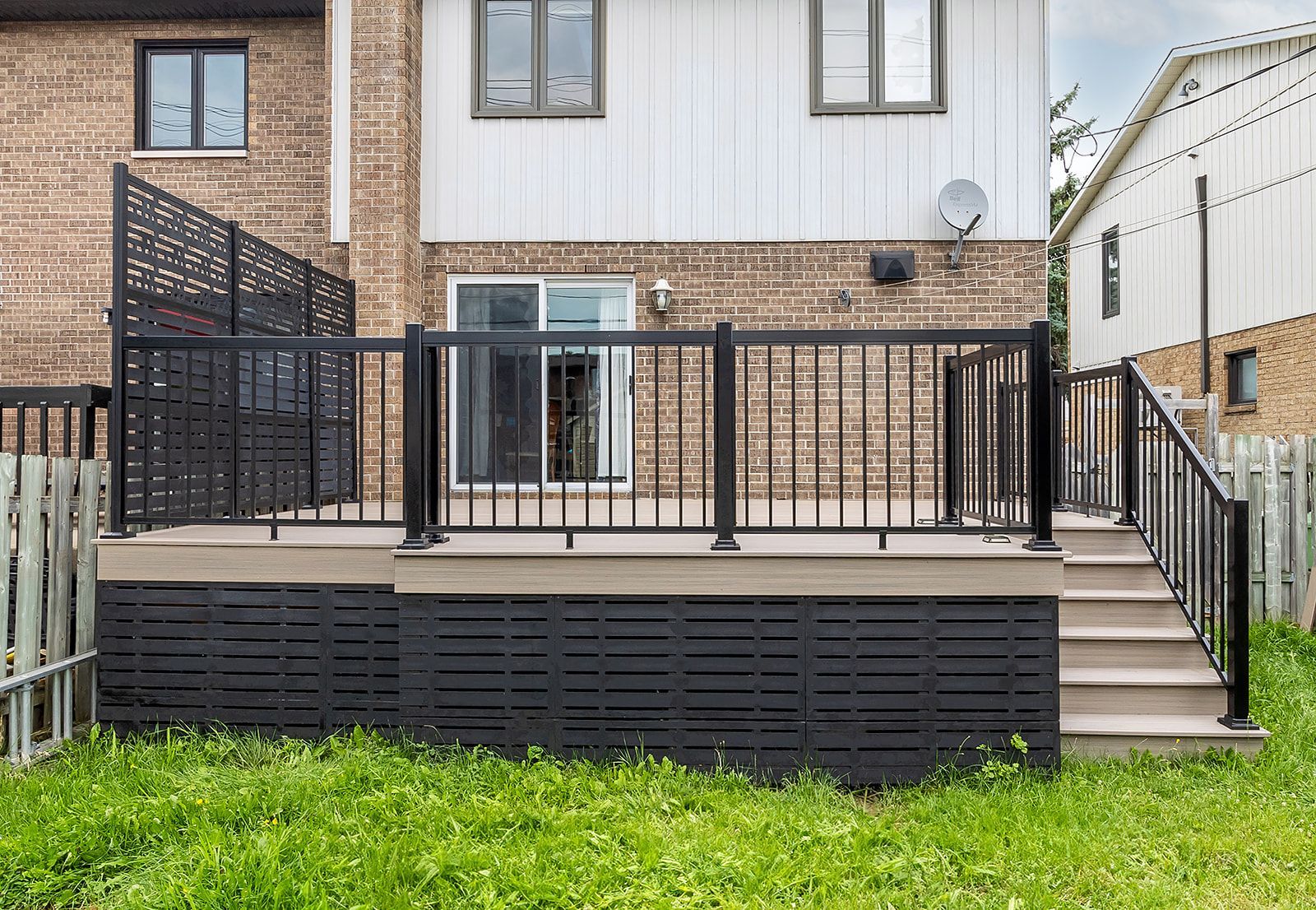 Terrasse noire avec rambarde et escalier, devant une maison blanche en briques, avec de l'herbe verte au premier plan.
