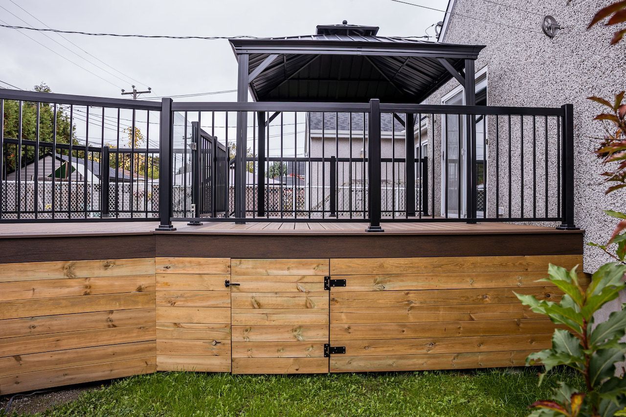 Terrasse avec rambarde noire, base en bois et kiosque à l'arrière-plan. Herbe et plantes au premier plan.
