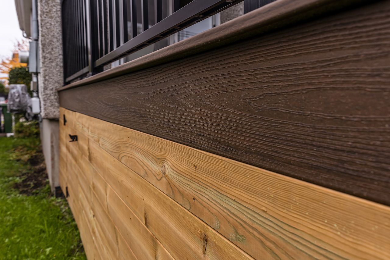 Gros plan sur une terrasse avec bardage en bois, finitions marron et rambarde noire. On aperçoit l'herbe.