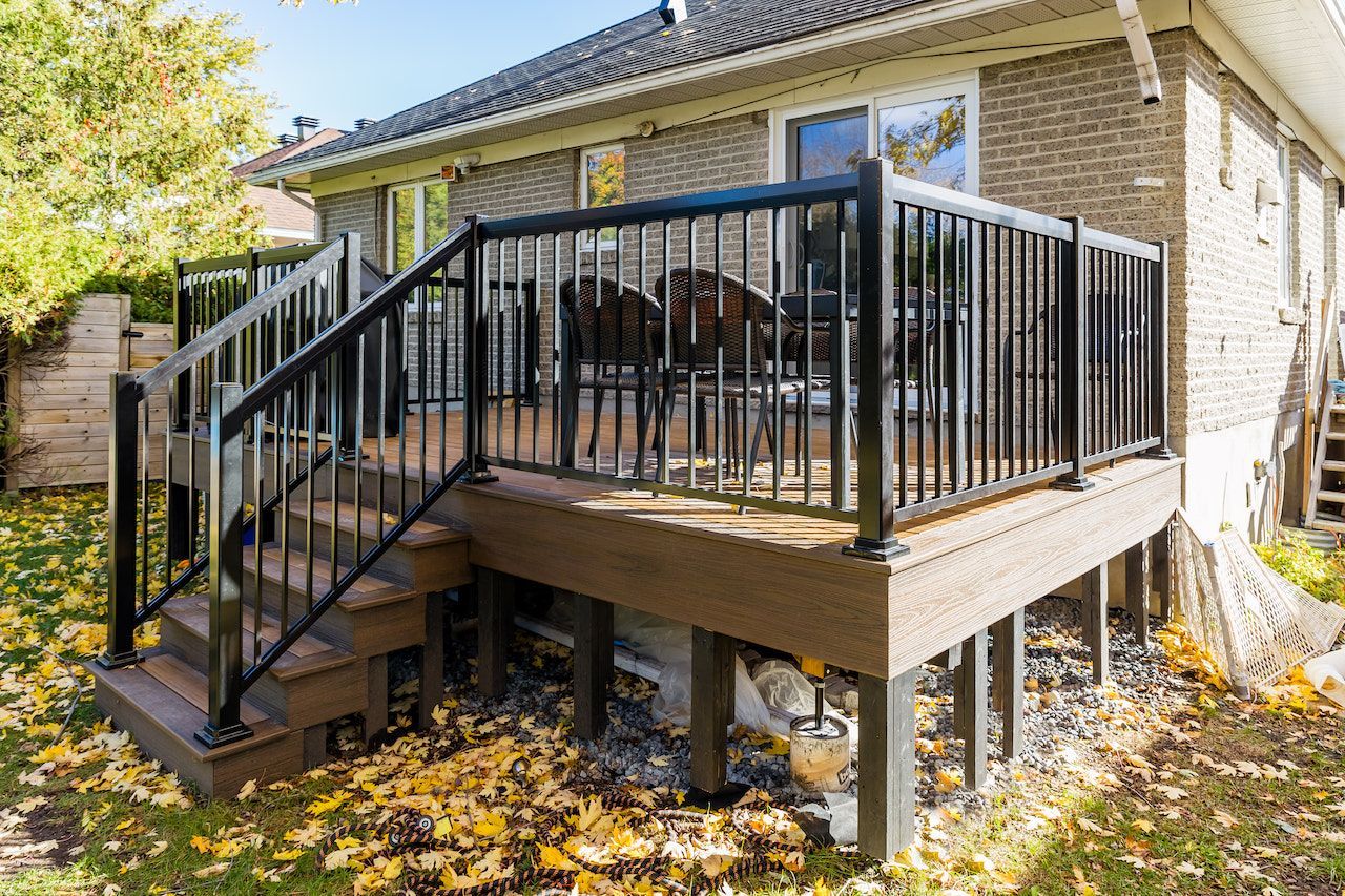 Terrasse avec rambarde noire, revêtement en composite et marches adossée à une maison en briques. Chaises et table visibles sur la terrasse.