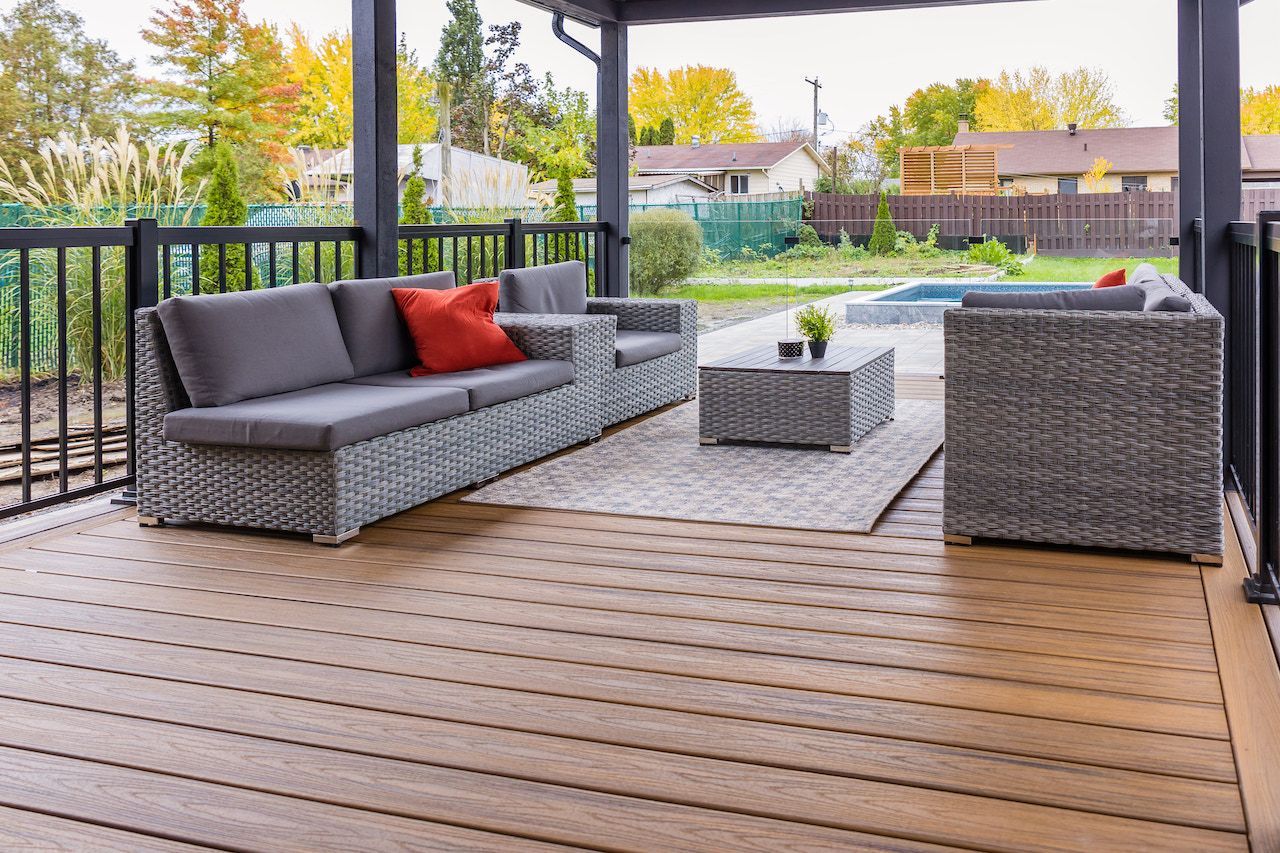 Terrasse extérieure avec mobilier en osier gris sur une terrasse en bois, donnant sur un jardin avec piscine.