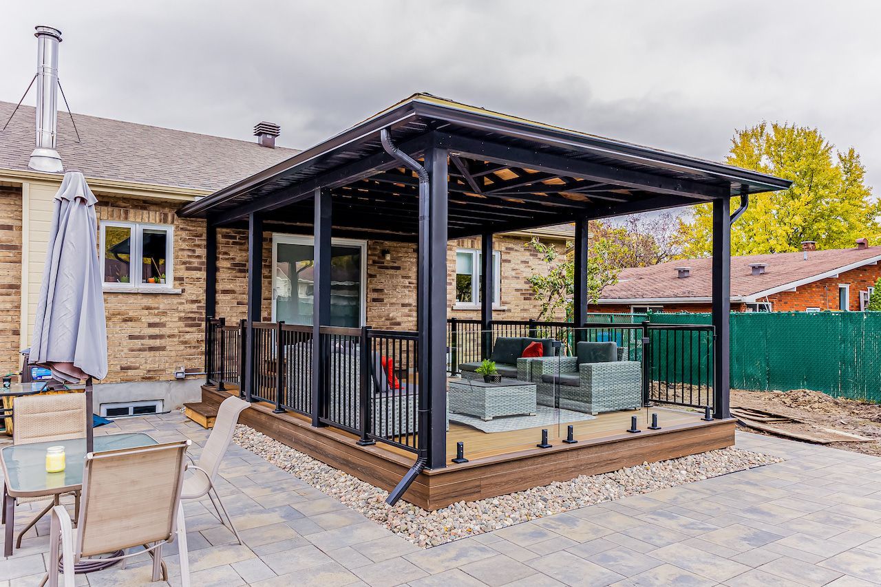 Auvent de patio à structure noire fixé à une maison en briques, au-dessus d'une terrasse avec mobilier de jardin.