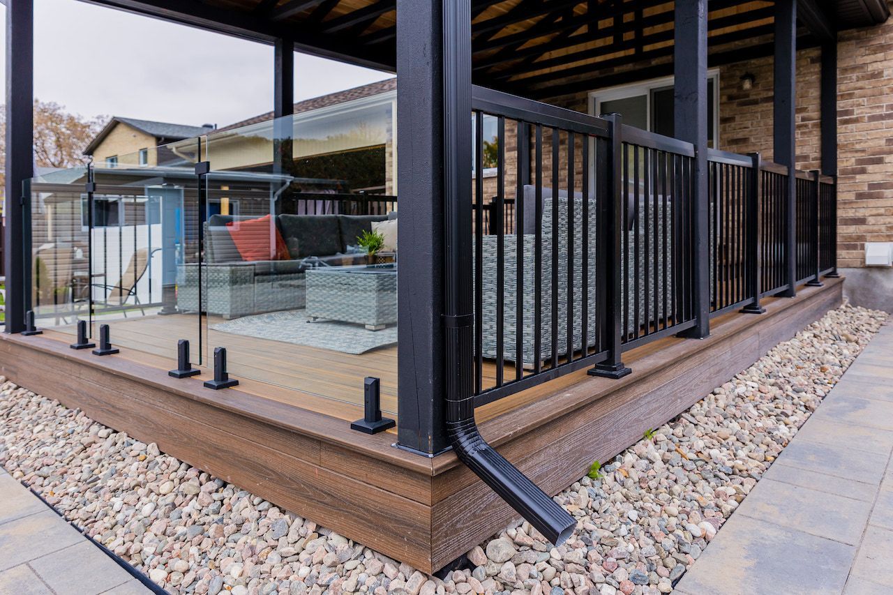 Terrasse extérieure couverte avec balustrades en verre et métal noir, et terrasse en composite marron, à côté d'un chemin en pierre.