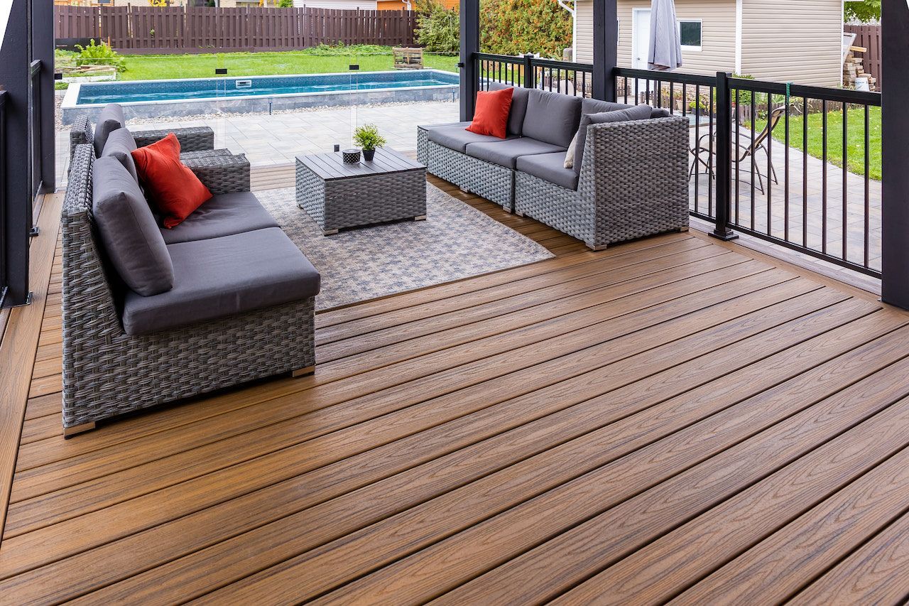 Terrasse en composite avec mobilier de jardin : canapés gris, coussins rouges, table basse et piscine en arrière-plan.