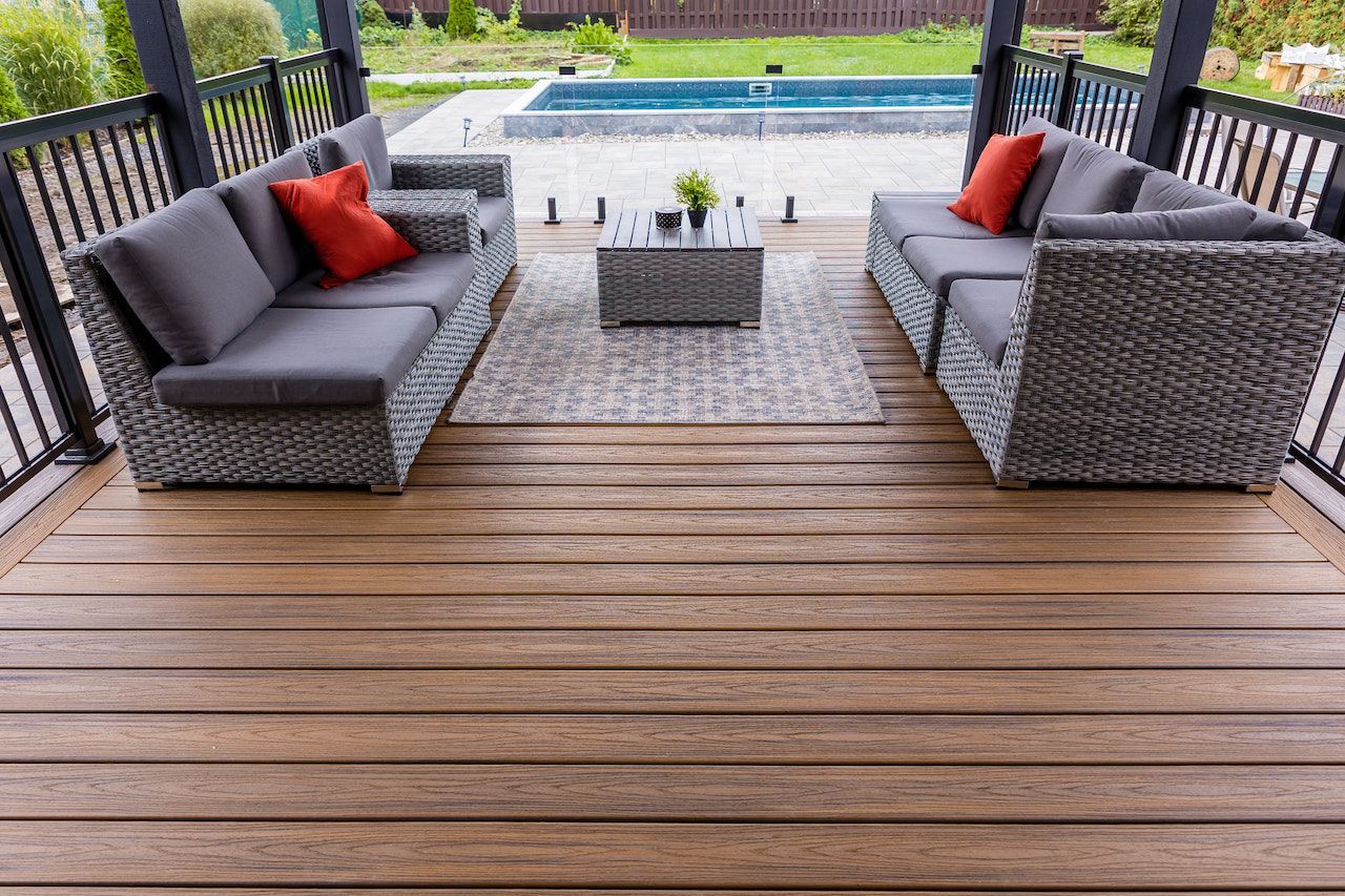 Terrasse extérieure avec canapés gris, coussins rouges, table basse, tapis et piscine en arrière-plan.