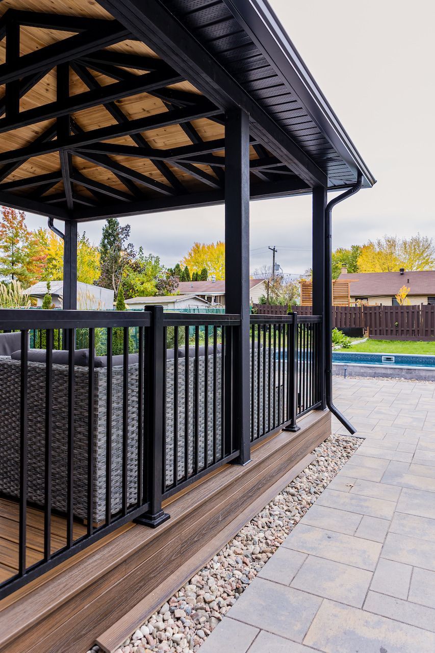 Gazebo noir avec rambarde et mobilier de jardin sur une terrasse en pierre donnant sur un jardin avec piscine.
