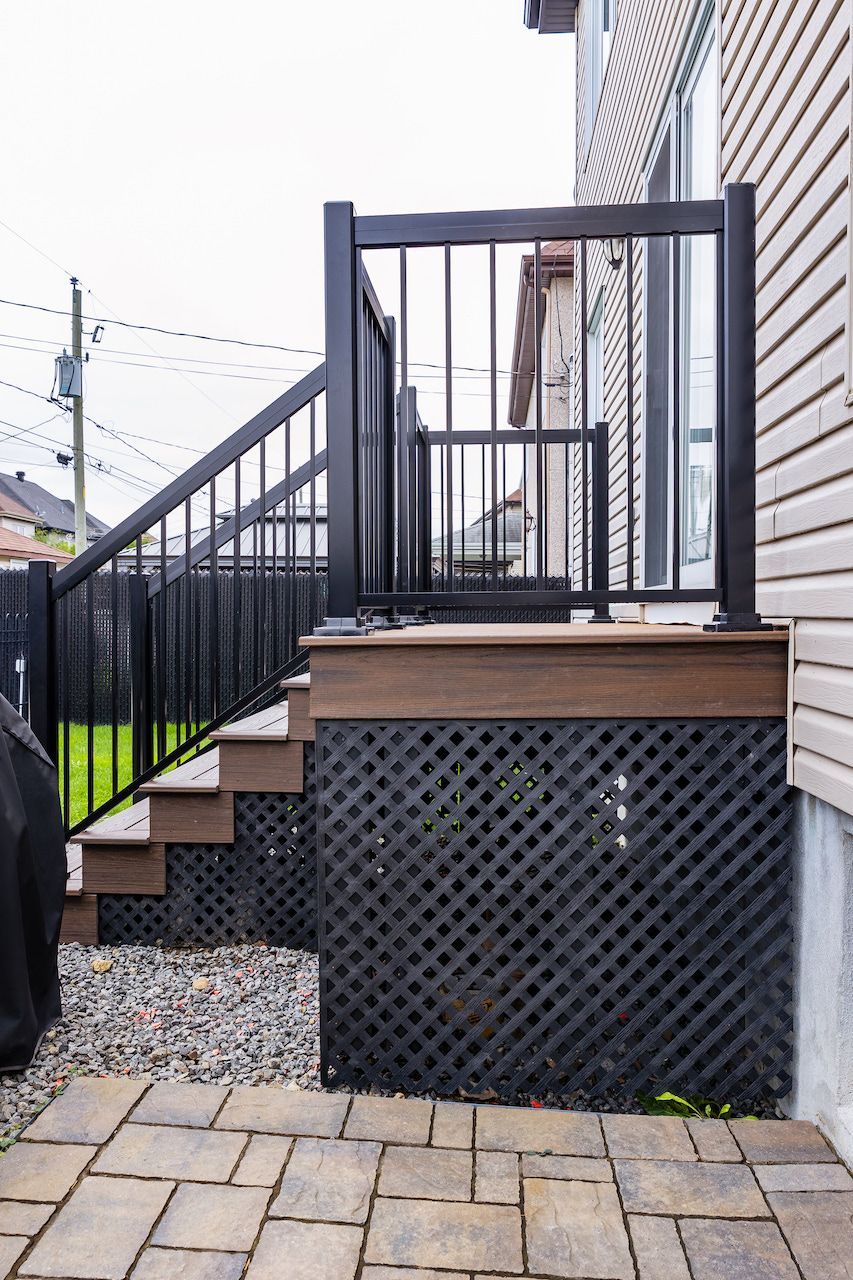 Rambarde et marches noires menant à une terrasse en treillis, à côté d'une maison beige avec un patio en briques.