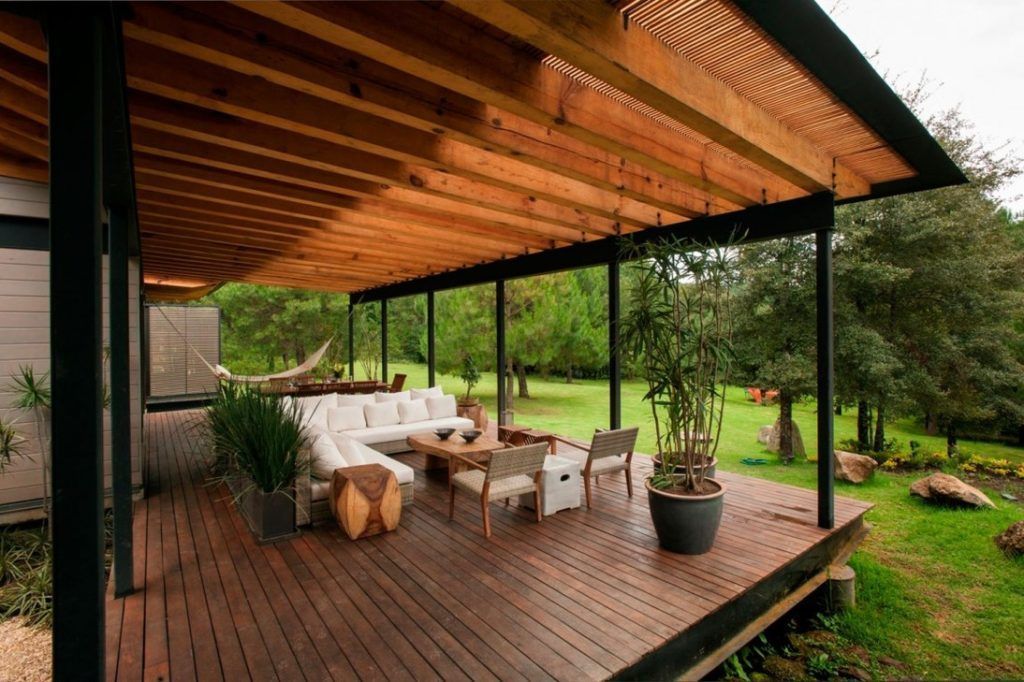 Terrasse extérieure couverte avec sièges, donnant sur une pelouse verdoyante et des arbres. Poutres et platelage en bois.