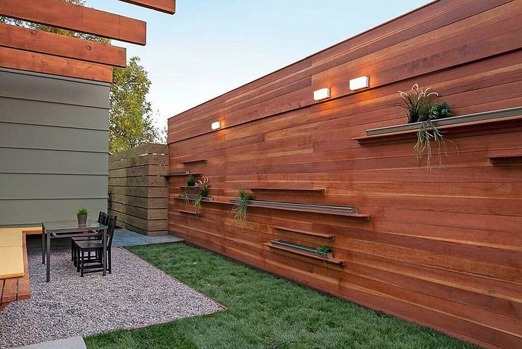 Clôture en bois avec étagères intégrées pour plantes, le long d'une cour gazonnée avec petite terrasse, table et chaises.