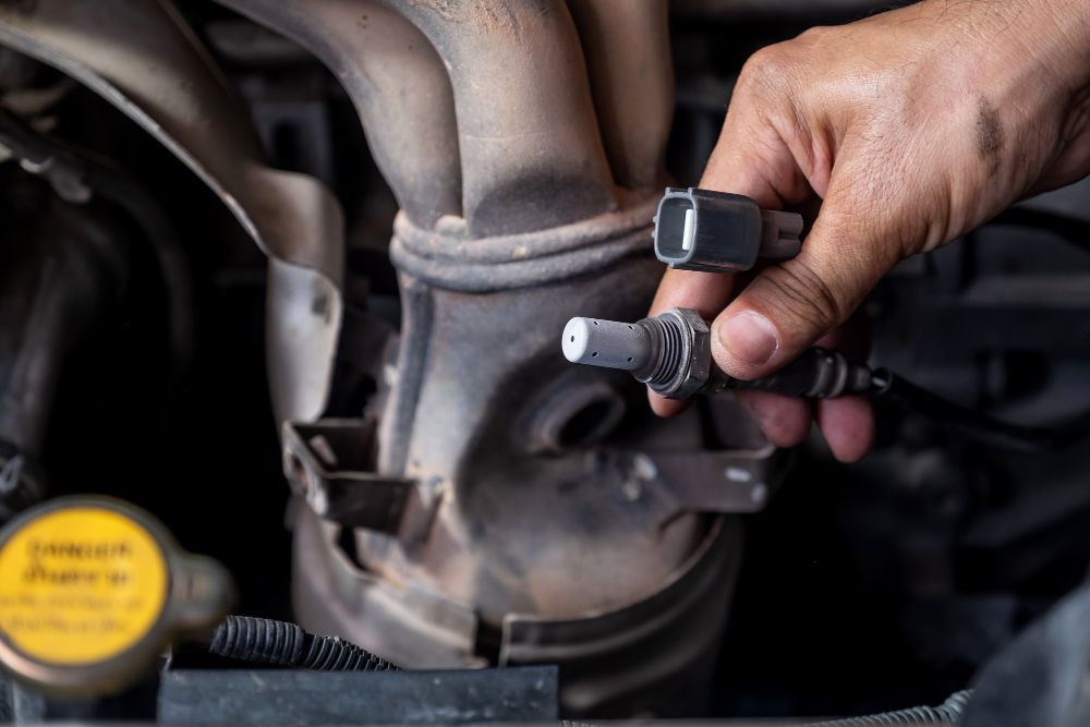 Hand holding oxygen sensor near car exhaust manifold.