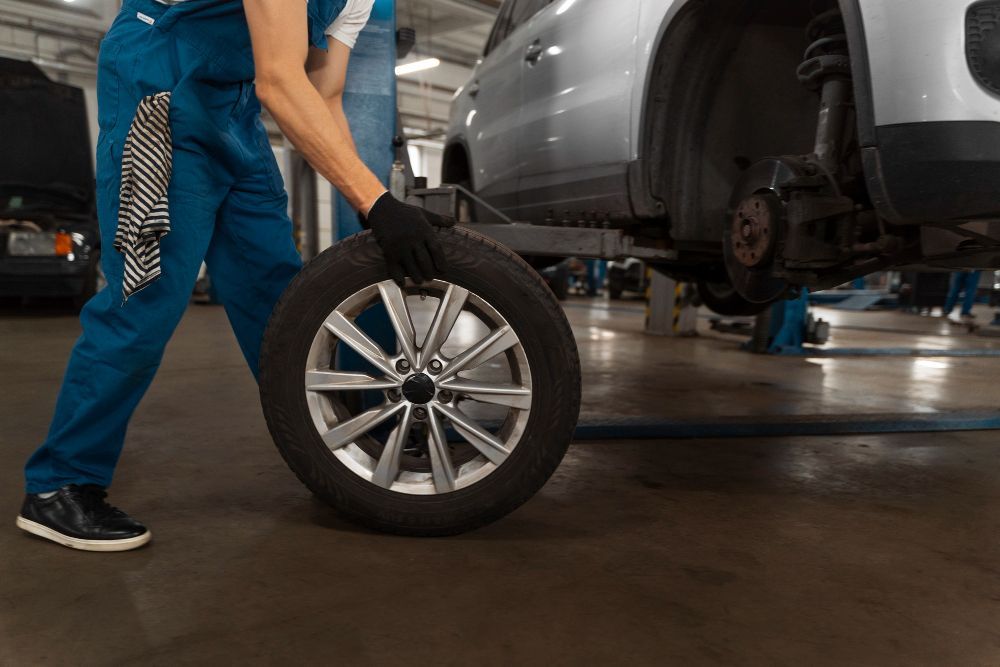 Mechanic in blue jumpsuit and gloves rolling a tire at a car repair shop.