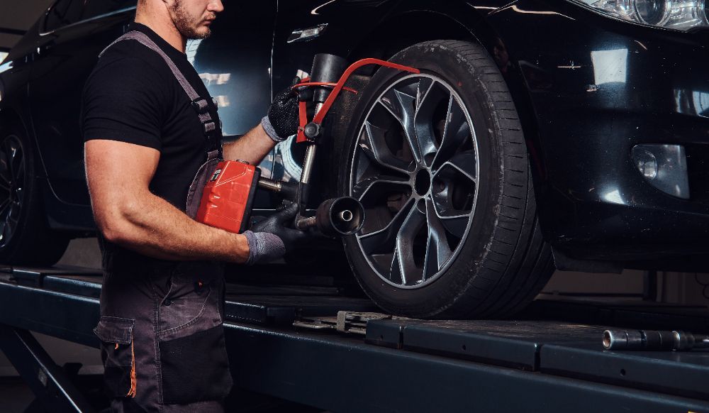 Mechanic balancing a black car tire on a lift, wearing gloves and holding a red tool.